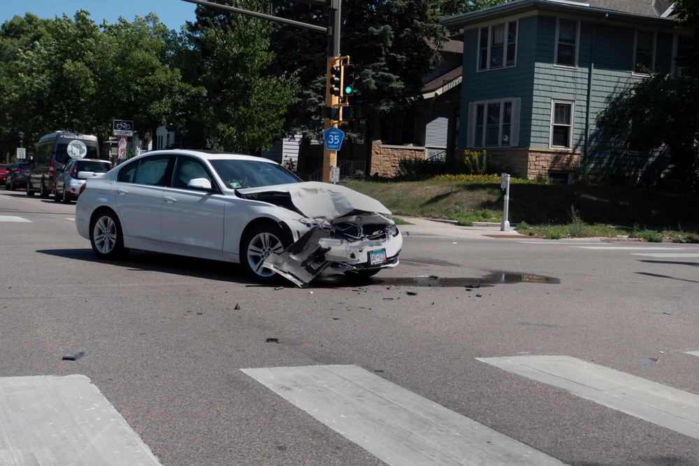 White sedan with severe front-end damage in the middle of an intersection after a car crash, with debris scattered on the road.