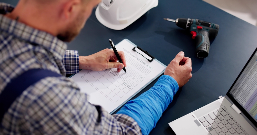Construction worker filling out a health insurance claim form at a desk, with a laptop, hard hat, and power drill nearby.